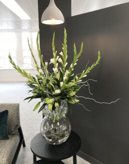 Photograph of lily bud heads inside a clear, round stacked vase, with white gladioli and white twigs, on a black table. Beside the table is a grey sofa with emerald velvet textured cushion. Corporate florist, event flowers, office flowers, sustainable florist, Central London florist, Local florist, Soho Florist, Contemporary Floral Display. Weekly Office Flowers; Weekly Flowers; Weekly Contract Flowers; Reception Desk Flowers; Office Reception Flowers