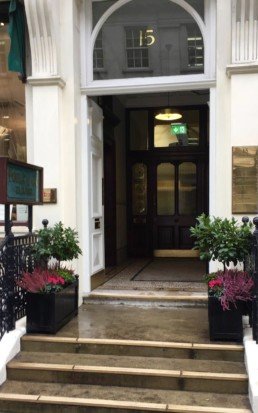 Photograph of vibrant winter plant maintenance for Savile Row, London, with pink Heather, hot pink Cyclamen and structured bay trees, in troughs either side of an open doorway. Mayfair; Luxury London; Soho Florist; Contract planting; plant Maintenance; landscaping; Corporate plants